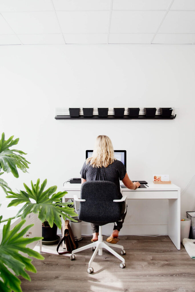 A woman sits at a desk in an office surrounded by plants.