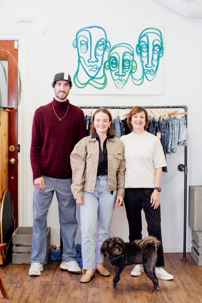 Three people smiling at the camera in front of their clothing racks in a consignment shop in Victoria, BC.