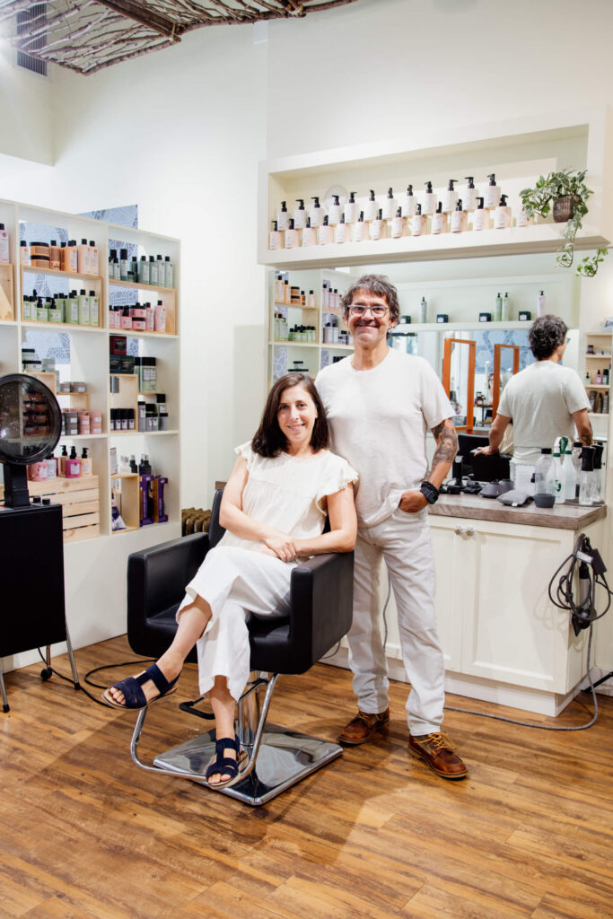 A woman in a hair salon sits in chair with a hairdresser standing beside her.