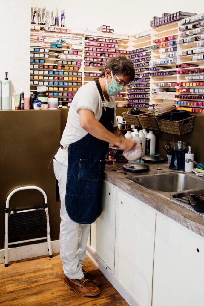 A hairdresser wearing a mask mixes hair colour over a sink.